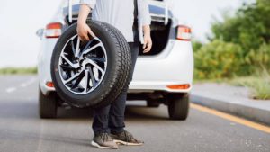 A person standing on the side of a highway next to a white car with its trunk open, holding a spare tire with a black and silver alloy wheel rim, illustrating a vehicle breakdown or puncture repair situation.