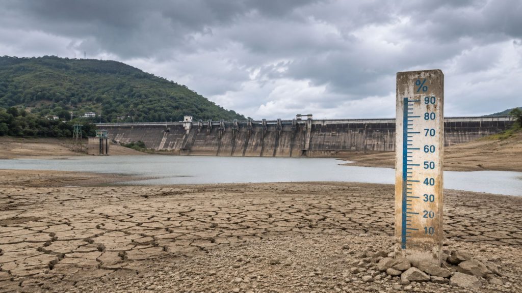 A drought-stricken reservoir near Pune with cracked, dry lakebed exposed in the foreground, a low water level visible behind it, and a large concrete dam stretching across the background under dark monsoon clouds. A weathered water-level gauge stands prominently beside the shrinking reservoir, illustrating critically low water storage as Pune awaits the delayed monsoon rains.