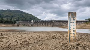 A drought-stricken reservoir near Pune with cracked, dry lakebed exposed in the foreground, a low water level visible behind it, and a large concrete dam stretching across the background under dark monsoon clouds. A weathered water-level gauge stands prominently beside the shrinking reservoir, illustrating critically low water storage as Pune awaits the delayed monsoon rains.