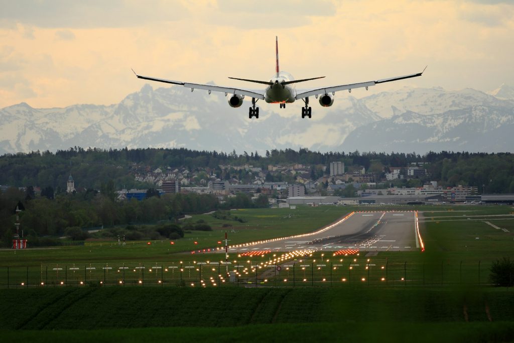 The shot of an aircraft from behind, coming in to land at a well-illuminated runway.