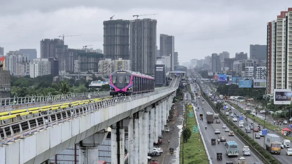 A Metro Line 3 train runs on the elevated Hinjawadi-Shivajinagar corridor above the Mumbai-Bengaluru Highway in Pune, with high-rise buildings, ongoing construction projects, and traffic visible below under cloudy monsoon skies.