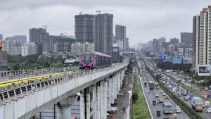 A Metro Line 3 train runs on the elevated Hinjawadi-Shivajinagar corridor above the Mumbai-Bengaluru Highway in Pune, with high-rise buildings, ongoing construction projects, and traffic visible below under cloudy monsoon skies.