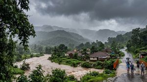 A heavy monsoon downpour over a rural village in the Maharashtra ghats, featuring a swollen, muddy river flowing rapidly next to houses with tiled roofs. Local residents carrying umbrellas and wearing raincoats walk along a wet, winding road under dark, stormy skies and misty green mountains.