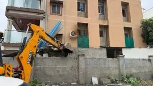 Pune News: A yellow Escorts JCB excavator demolishes an unauthorized concrete wall and temporary structure in front of a multi-story residential building during a Pune Municipal Corporation (PMC) demolition drive.